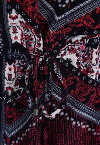 Gemustertes Kleid aus Stoff mit roten, weißen und schwarzen Designs. Verfügt über eine gebundene Taille und detaillierte grafische Muster in unterschiedlichen Farben und Texturen.