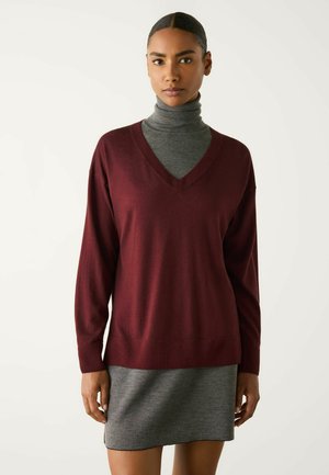 Kvinde med mørkt hår i en lav knold iført en grå rullekræset kjole og en løs vinrød V-hals sweater, stående mod en ensartet lys baggrund.