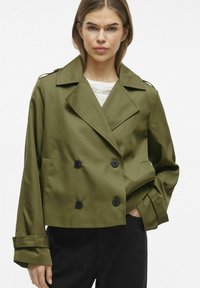 Divkāršs olīvzaļš žakets ar lieliem melniem pogām, plašām atlokām un saīsinātām piedurknēm, izgatavots no gludas auduma, ar sānu kabatām.