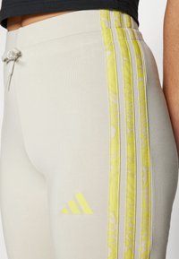 Svetlo sive legginge z rumenimi črtami in adidas logotipom. Narejene so iz elastičnega blaga, ki ima gladko teksturo in pas na vrvico.