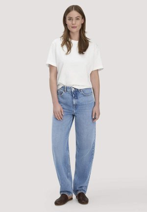 Vrouw staand, gekleed in een wit T-shirt met korte mouwen, lichtblauwe high-waisted jeans en bruine instappers, voor een effen achtergrond.