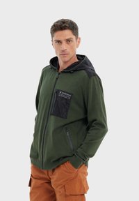 Haut à capuche zippé vert avec une capuche noire et des accents noirs sur les épaules, comportant une poche poitrine noire et des poches zippées sur les côtés. Texture lisse.