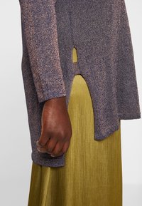 Zbliżenie ciemnoskórej dłoni obok fioletowego swetra z dzianiny z bocznym rozcięciem na oliwkowozielonej plisowanej spódnicy.