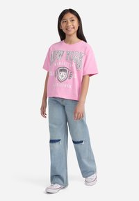 Tricou din bumbac roz cu grafică "NEW YORK", asortat cu blugilargi de culoare albastră deschisă, cu pete albastre, și adidași albi.