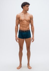 Γυμνασμένος νέος άνδρας με σκουρόχρωμα σγουρά μαλλιά στέκεται ξυπόλητος φορώντας σκούρα πετρόλ μποξεράκια με μαύρη μέση και γεωμετρικό σχέδιο, κοιτάζοντας προς το πλάι.