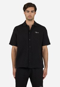 Black short-sleeve shirt with a textured diamond pattern, featuring a button-front closure and a logo embroidered on the left chest.
