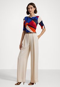 Top de patrón colorido con formas azules, rojas y beiges, combinado con pantalones beiges de cintura alta, acentuado por un cinturón marrón y tacones.