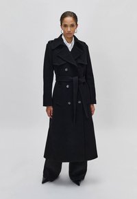 Mulher em pé, vestindo um longo casaco trench preto duplo com cinto, calças pretas e sapatos pretos de bico em um fundo cinza liso.