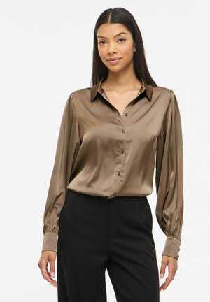 Mujer con blusa marrón brillante de manga larga y botones, y pantalones negros, de pie frente a un fondo claro y liso.