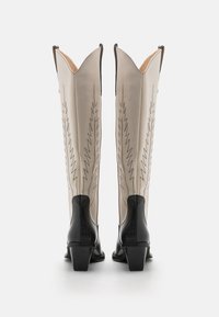 Knee-high cowboy saapad kreemjas beezis nahas koos mustade aktsentidega, teravate tippudega ja keeruka sakilise õmblusega külgedel.