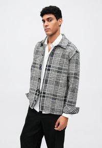 Καπιτονέ σακάκι σε γκρι και ναυτικό χρώμα με κλασικό γιακά, κουμπί και μαλακή υφή. Φοριέται πάνω από ένα λευκό πουκάμισο και μαύρο παντελόνι.