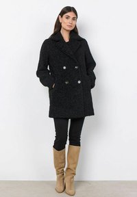 Abrigo negro de doble botonadura con una superficie texturizada y suave, dos bolsillos frontales y botones plateados. Combinado con pantalones negros y botas beiges.