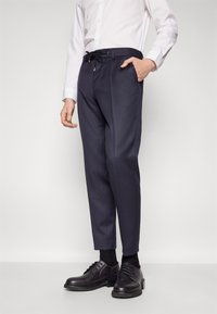 Navy trousers with a tapered fit, featuring a drawstring and belt loops. Worn with a white button-up shirt and black leather shoes.