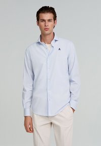 Helleblaue gestreifte Button-Down-Bluse mit langen Ärmeln, die ein kleines dunkelblaues Logo auf der Brust trägt. Hergestellt aus einem glatten, leichten Stoff.