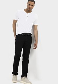 Biała, krótko rękawowa koszula henley z trzema guzikami, zestawiona z czarnymi dżinsami i jasnobeżowymi trampkami. Model stoi na jednolitym tle.