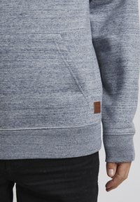 Sudadera gris de tejido suave y texturizado con un bolsillo canguro y puños de canalé. Presenta una pequeña etiqueta de cuero marrón en el dobladillo.