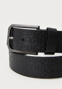 Black leather belt featuring embossed "HUGO" logo, with a shiny black metal buckle and smooth texture. Accents include a single prong design.