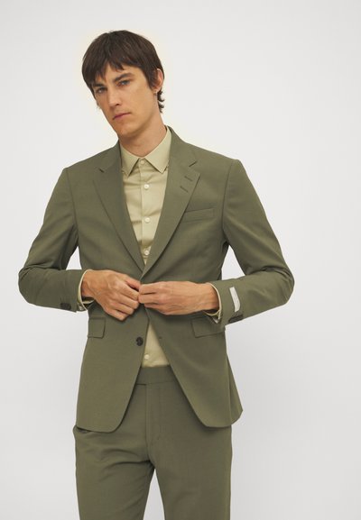 Olivgrüner Anzug im Slim-Fit mit einreihigem Design, Spitzenrevers und passender Hose. Darüber getragen wird ein beiges Hemd mit Knöpfen.