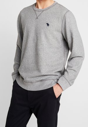 Mann trägt ein hellgraues Langarm-Sweatshirt mit kleinem dunklem Tierlogo auf der Brust und schwarze Hosen, die Hände in den Taschen.