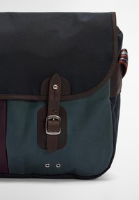 Mala mensageiro em lona e couro com aba escura, fivela de metal, bolso frontal em teal e laço de tecido multicolorido de um lado.