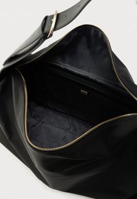 Μαύρη σύνθετη τσάντα χειρός με λεία υφή, φερμουάρ σε χρυσό τόνο και ευρύχωρο εσωτερικό με διακριτικό σχέδιο λογότυπου "GUESS".