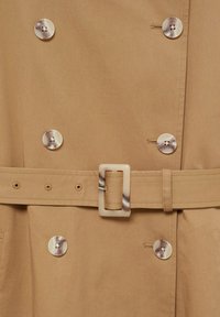 Trench-coat beige présentant une texture lisse, un design croisé à double boutonnage, de gros boutons et une ceinture assortie avec une boucle rectangulaire.