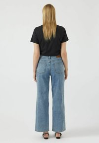 Jeans de pernera ancha en un azul claro con una cintura alta, dos bolsillos traseros y una etiqueta de cuero marrón en la cinturilla, combinados con una camiseta negra.
