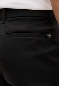 Κοντινό πλάνο πίσω τσέπης μαύρου παντελονιού με ετικέτα λογότυπου Puma, φορεμένο με λευκό πουκάμισο μέσα.