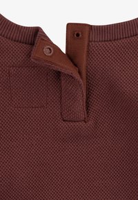 Maglione marrone scuro lavorato a maglia con una trama testurizzata, dotato di un colletto con bottoni e una piccola toppa vicino alla spalla.