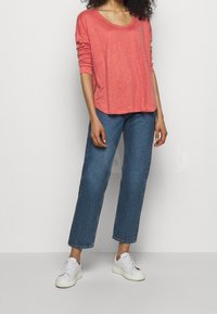 Korallin värinen pitkähihainen paita, jossa on avara kaula-aukko, yhdistettynä löysiin sinisiin farkkuihin. Valkoiset tennarit täydentävät asukokonaisuuden. Yksinkertainen muotoilu, pehmeä rakenne.