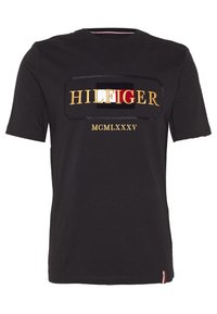 Svart kortärmad T-shirt med "HILFIGER" och "MCMLXXXV" broderat i guld, med en rektangulär logobakgrund i röd, vit och svart.