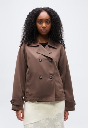 Kobieta o kręconych włosach, ubrana w brązową dwurzędową marynarkę i białą teksturowaną spódnicę, stojąca na tle jasnego, jednolitego tła.