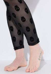 Leggings neri trasparenti con un motivo a teschi su tutta la superficie. Il tessuto ha una texture liscia e una lunghezza fino alla caviglia aderente con orlo a bande.