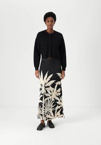 Zwarte cropped cardigan met een knoopsluiting, gecombineerd met een vloer-lange rok met grote witte botanische prints. Zwarte instapschoenen.