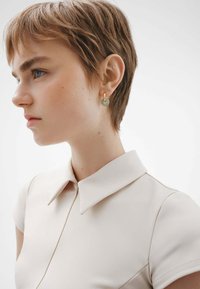 Camisa color beige con cuello y mangas cortas, de textura suave, que presenta un cierre oculto. Aros de plata y oro adornan la oreja de la modelo.