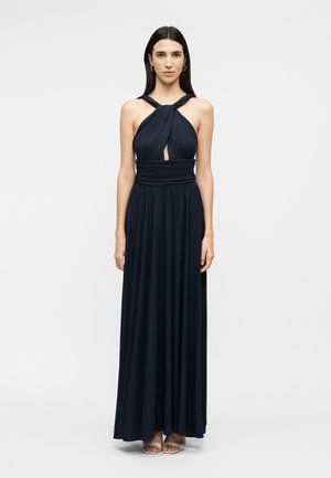 Γυναίκα με μακριά σκουρόχρωμα μαλλιά, που φοράει μαύρο maxi φόρεμα χωρίς μανίκια με στριφτό ντεκολτέ και λεπτομέρεια κοψίματος, στέκεται μπροστά σε λευκό φόντο.