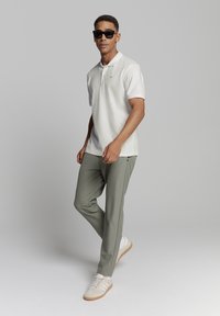 Witte poloshirt, olijfgroene broek en lichte sneakers. Zonnebril gedragen, wat een moderne casual outfit aanpraat. Glad materiaal en op maat gemaakte pasvorm.