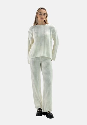 Gestreifter Strickpullover und passende Hose in Off-White, mit lockerer Passform, langen Ärmeln und strukturiertem Stoff. Schwarze klobige Schuhe als Fußbekleidung.