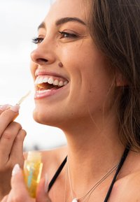 Durchsichtiger Lipgloss in einem gelben Behälter, aufgetragen von einer Person mit langen, dunklen Haaren, die ein Lächeln und weiße Zähne zeigt.