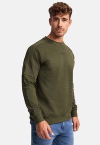 Sudadera verde oliva con cuello redondo, puños acanalados y un tejido texturizado. Presenta un pequeño logo en el pecho y una etiqueta lateral.