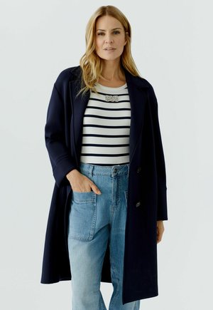 Femme blonde portant un manteau bleu marine, un haut blanc à rayures bleu marine avec une broche en strass, et un jean en denim bleu clair avec des poches à patch.