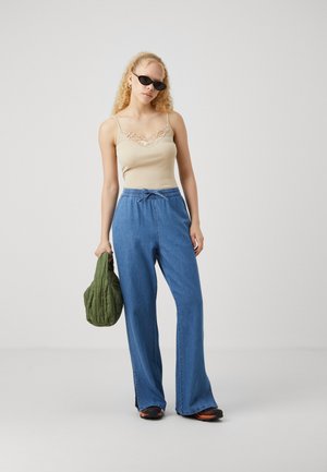 Top in beige con pizzo, pantaloni in denim a gamba larga blu e borsa trapuntata verde. La modella indossa occhiali da sole neri e calzature nere.