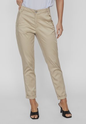 Woman wearing beige rolled-up pants with front pockets and black open-toe heeled mules, standing against a plain background.