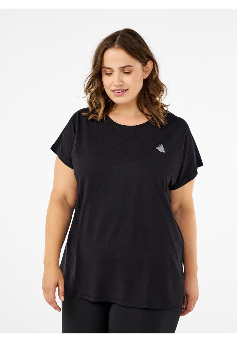 Femme aux cheveux bruns ondulés portant un t-shirt noir ample avec un petit logo blanc sur la poitrine, debout devant un fond blanc.