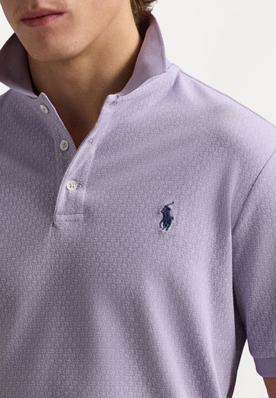 Lilla polo särk, millel on tekstuuriga kangas, kahe nööbiga kaelus ja väike tumesinine logo aktsent rinnal. Lühikesed varrukad.