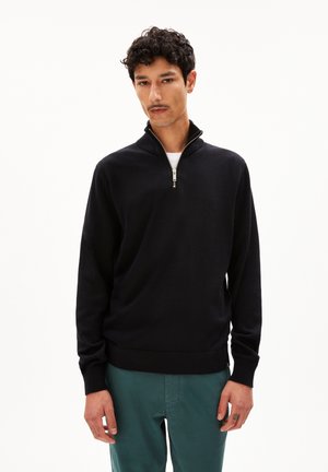 Schwarzer Pullover mit Reißverschlusskragen aus weichem Strickmaterial. Verfügt über gerippte Bündchen und Saum, getragen über einem weißen Shirt mit schlichtem Design.
