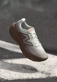 Baskets de course New Balance blanches et beiges avec semelle Fresh Foam flottant au-dessus d'un revêtement en asphalte avec des marquages routiers blancs.