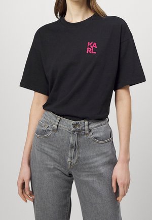 Frau trägt ein schwarzes T-Shirt mit pinkfarbenem "KARL"-Schriftzug auf der Brust und hoch taillierte, ausgewaschene graue Jeans vor einfachem Hintergrund.