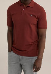Rödbrun polo-shirt i ett mjukt material, med en krage, korta ärmar, två knappar och en liten broderad logotyp på bröstet.