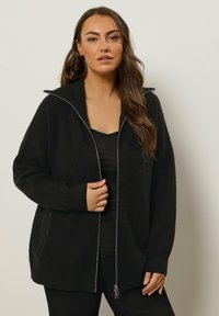 Czarny kardigan na zamek z prążkowaną teksturą, w oversize'owym fasonie i wysokim kołnierzem. Nosi się go na czarnym topie, ma srebrny zamek błyskawiczny i boczne kieszenie.